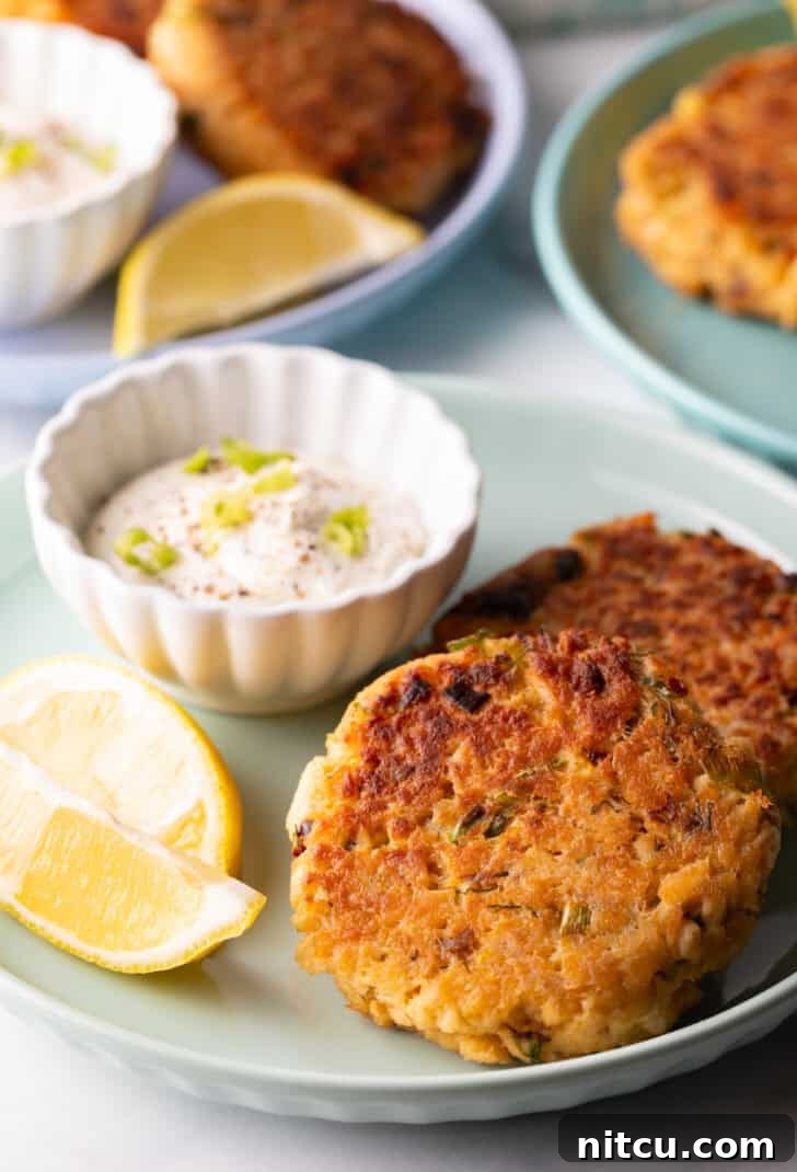 Two golden-brown salmon patties presented on a vibrant green plate, accompanied by a small white ramekin of creamy tartar sauce and two bright lemon wedges.