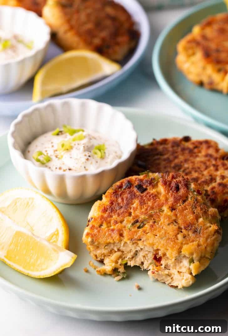 Two golden-brown salmon patties rest on a green plate, with a small white ramekin of tartar sauce and two lemon wedges. A noticeable bite has been taken from the patty closest to the camera, showcasing its flaky interior.