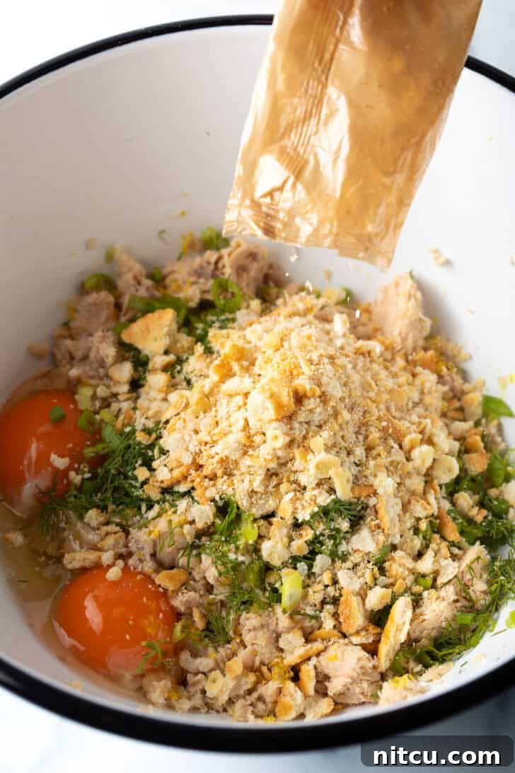 A large white mixing bowl containing flaked canned salmon, crushed crackers, vibrant fresh herbs, and cracked eggs, ready to be combined for salmon patties.
