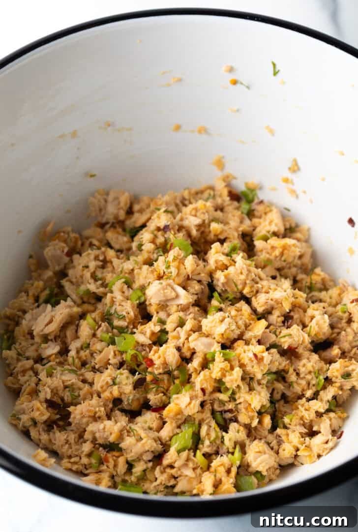 A close-up shot of flaked salmon combined with crushed crackers and freshly chopped herbs in a white mixing bowl, showcasing the texture before forming patties.