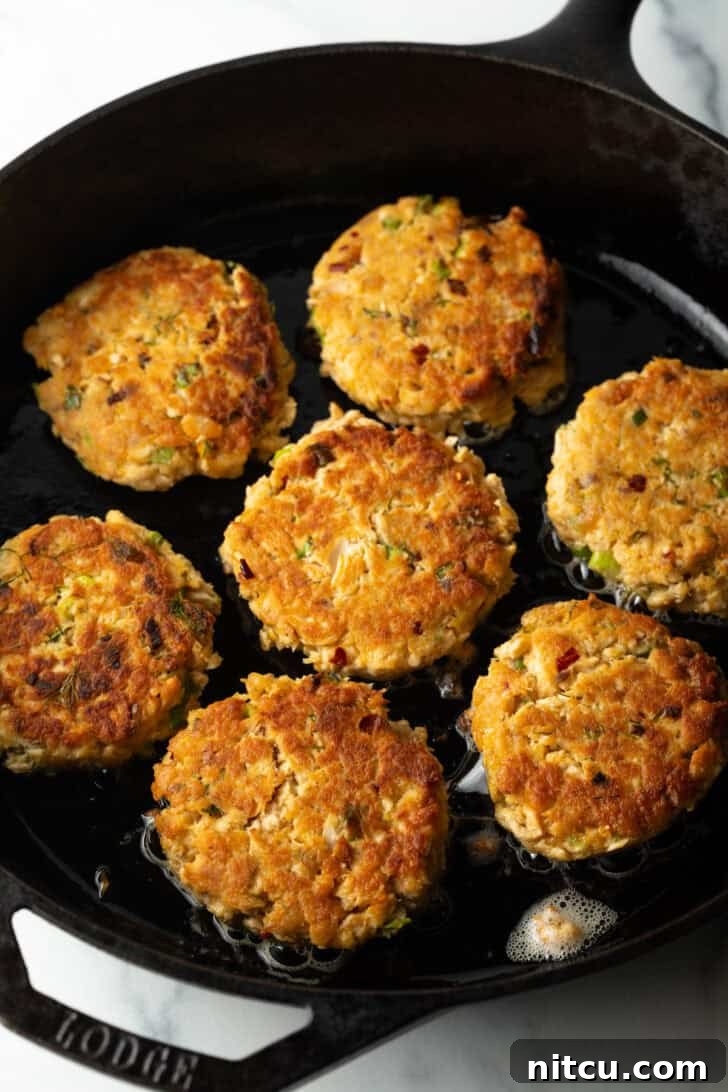 A top-down view captures seven salmon patties sizzling and browning beautifully in a cast iron skillet, illustrating the pan-frying process.