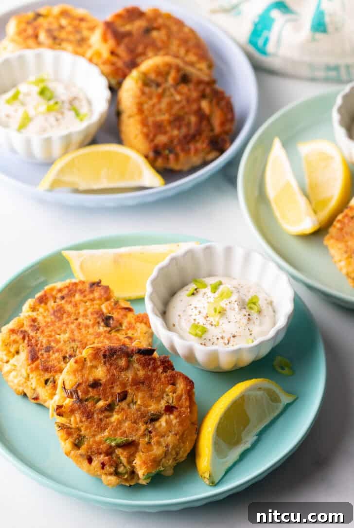 A beautifully arranged serving of old-fashioned salmon patties on a green plate, complete with a ramekin of creamy tartar sauce and fresh lemon wedges.