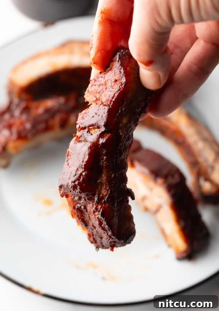 A hand holding a single, perfectly air-fried baby back rib, coated in shimmering barbecue sauce.