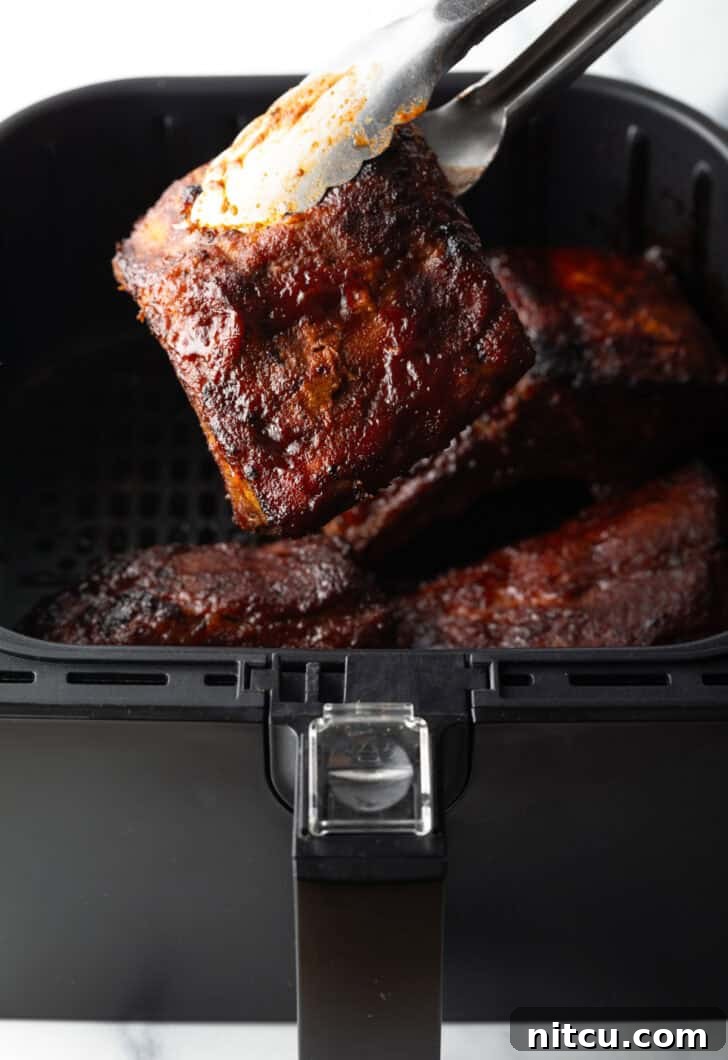 Tongs pulling a perfectly cooked, glazed rib from an air fryer basket, with another juicy rib held up to the camera.