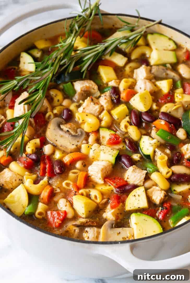 A large white pot overflowing with homemade chicken minestrone soup, garnished with fresh sprigs of rosemary, ready to be served.