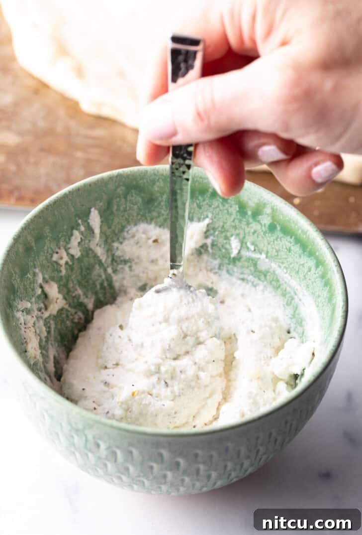 A hand stirring white sauce ingredients in a green bowl with a spoon.