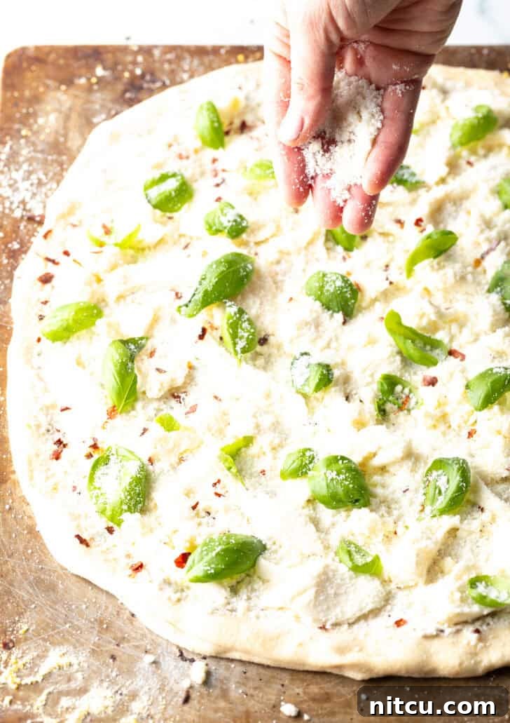 A hand adding shredded mozzarella cheese on top of the unbaked white pizza.