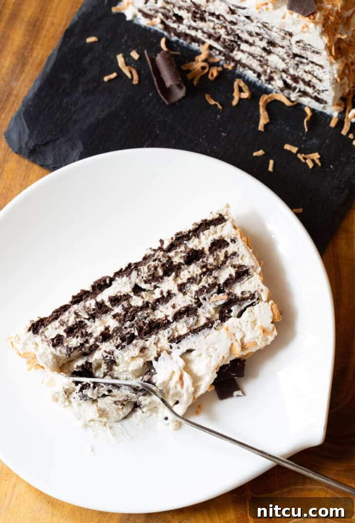 Slice of ice box cake with layers of chocolate graham crackers and Kahlua whipped cream, topped with toasted coconut and chocolate shavings on a white plate. A fork is taking a bite from the bottom of the slice.