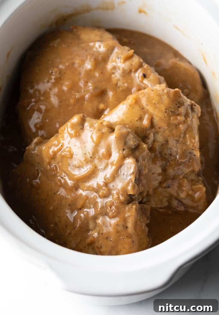 Three large bone-in pork chops perfectly cooked and submerged in a rich onion gravy within a white crock pot, steaming gently.