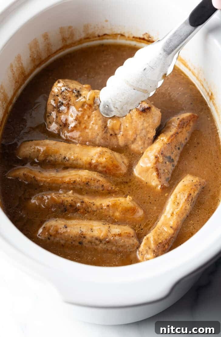 Tongs gently lifting a perfectly cooked pork chop from a white slow cooker, filled with other chops and rich onion gravy.