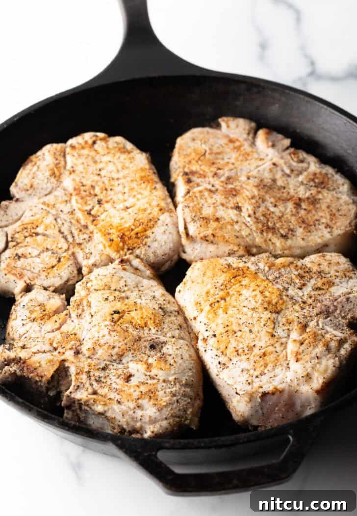 Four bone-in pork chops browning in a cast iron skillet, their edges caramelizing beautifully.