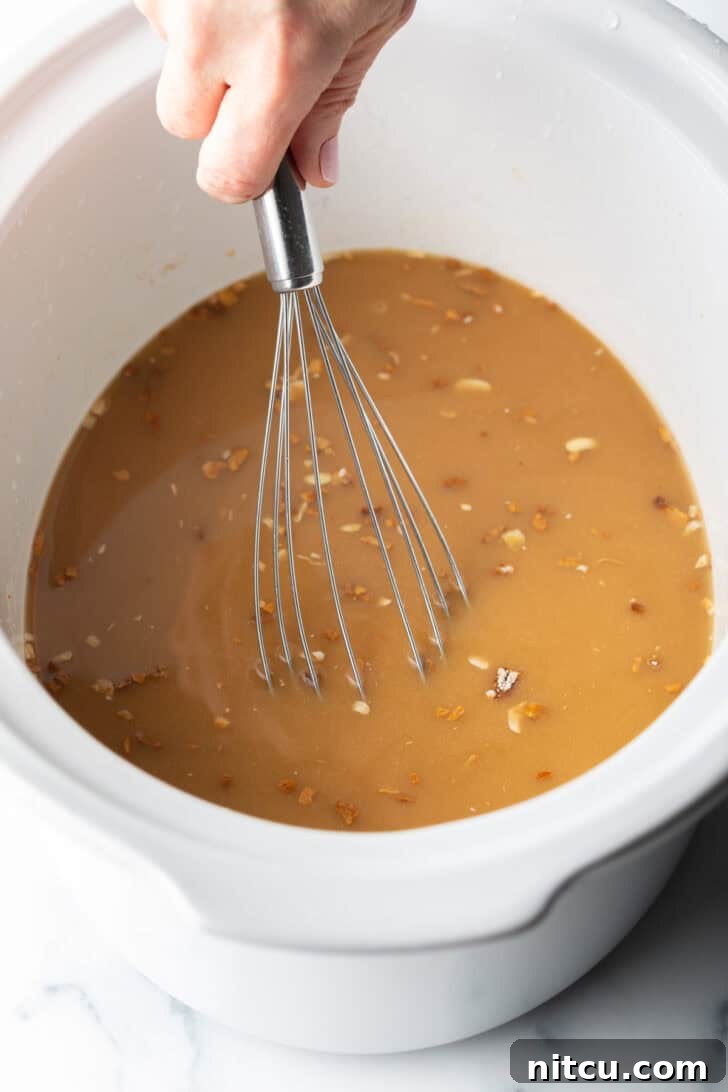 A hand using a whisk to thoroughly combine the brown onion gravy ingredients directly in a white slow cooker.
