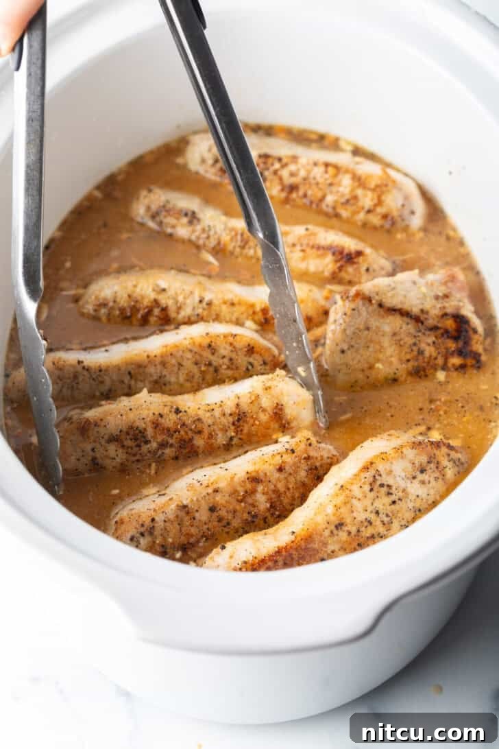 Tongs carefully placing a seared pork chop into a white slow cooker filled with simmering onion gravy.
