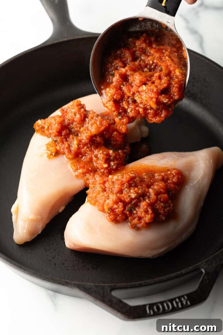 Adding tomato salsa from a measuring cup onto two raw chicken breasts in a cast iron skillet, beginning the cooking process.