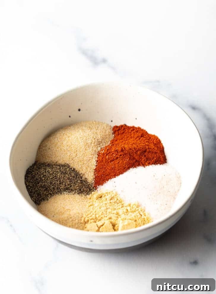 Separate, neatly arranged piles of smoked paprika, salt, onion powder, black pepper, garlic powder, ground mustard, and a small pinch of cayenne pepper in a white bowl, ready to be mixed.