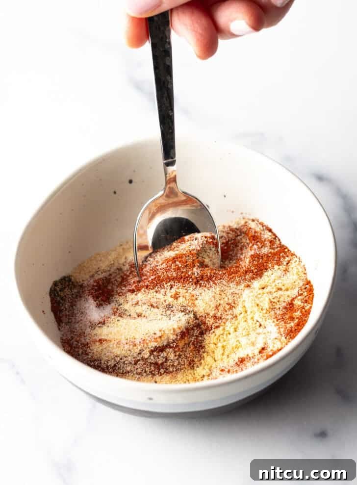 A hand stirring together various spices in a white bowl with a spoon, showing the blending process of the burger seasoning.
