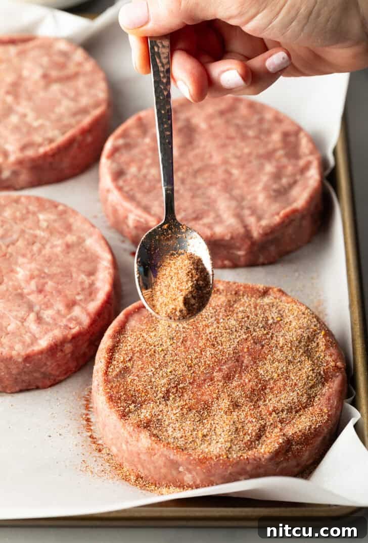 A metal spoon sprinkling a generous amount of homemade burger seasoning onto a raw hamburger patty.