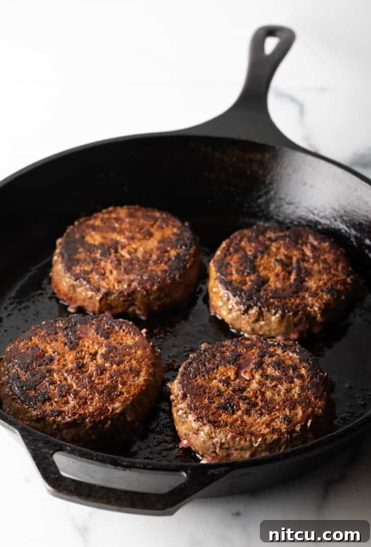 Four perfectly cooked hamburgers resting in a well-seasoned cast iron skillet, showcasing their delicious crust.