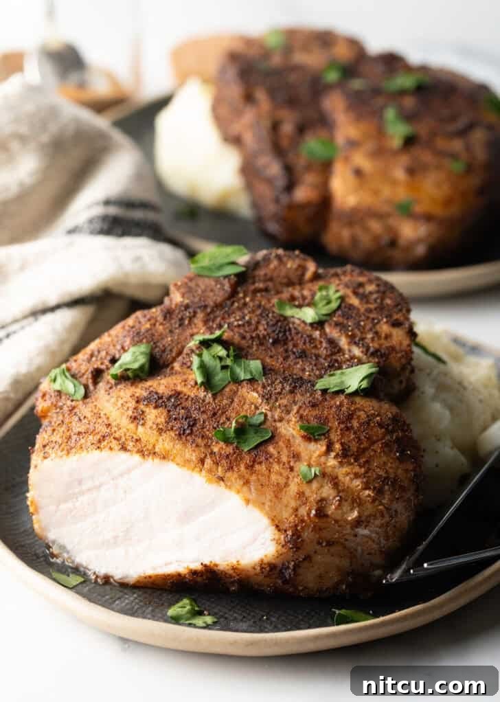 Browned seasoned bone-in pork chop on a plate with mashed potatoes, garnished with chopped parsley. The front piece of the chop has been cut off to show the juice meat inside.