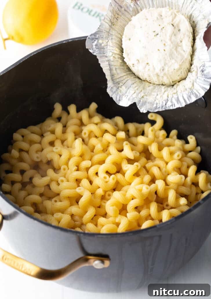 Adding creamy Boursin cheese directly from its wrapper into a pot of freshly cooked macaroni noodles.