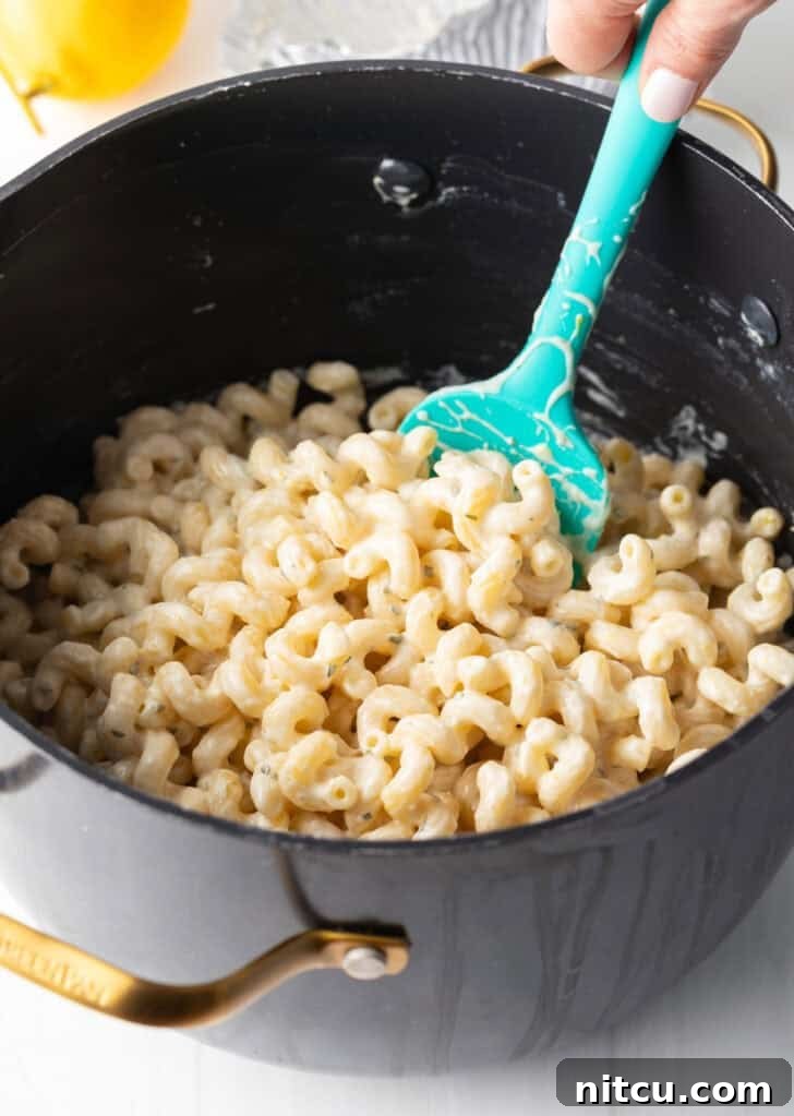 A hand stirring pasta and a rich cheese sauce in a large pot using a blue spatula, ensuring every noodle is coated.