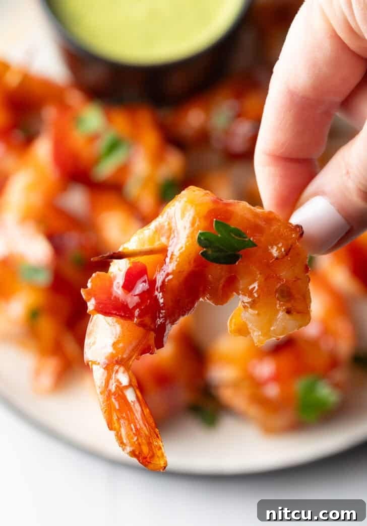 A hand gently holds a single, perfectly cooked bacon-wrapped shrimp close to the camera, showcasing its crispy texture and succulent appearance, ready for a bite.