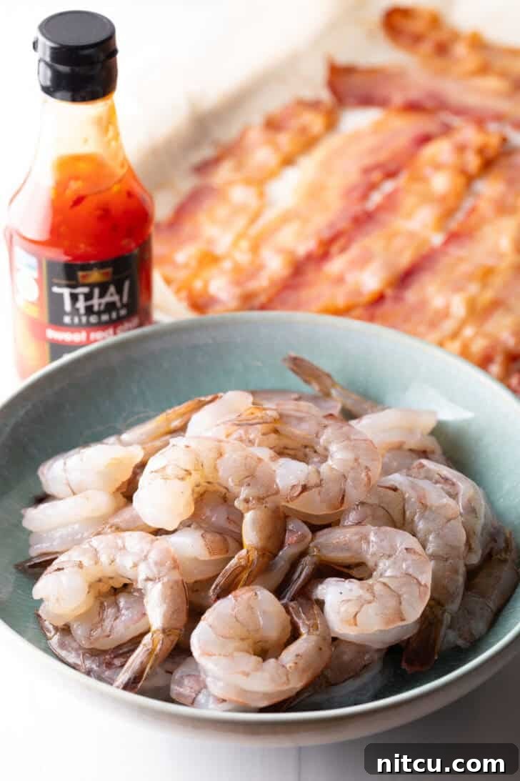 A bowl of raw, pink shrimp sits prominently on a kitchen counter, flanked by a glistening bottle of vibrant red Thai chili sauce. In the blurred background, a baking sheet already prepped with bacon hints at the recipe's next steps, creating a scene of culinary anticipation.