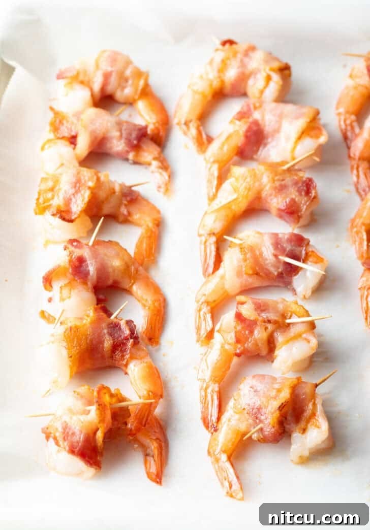 A dozen succulent shrimp are meticulously wrapped with strips of bacon, each held securely in place by a small wooden toothpick, arranged on a baking sheet, poised for their final bake.