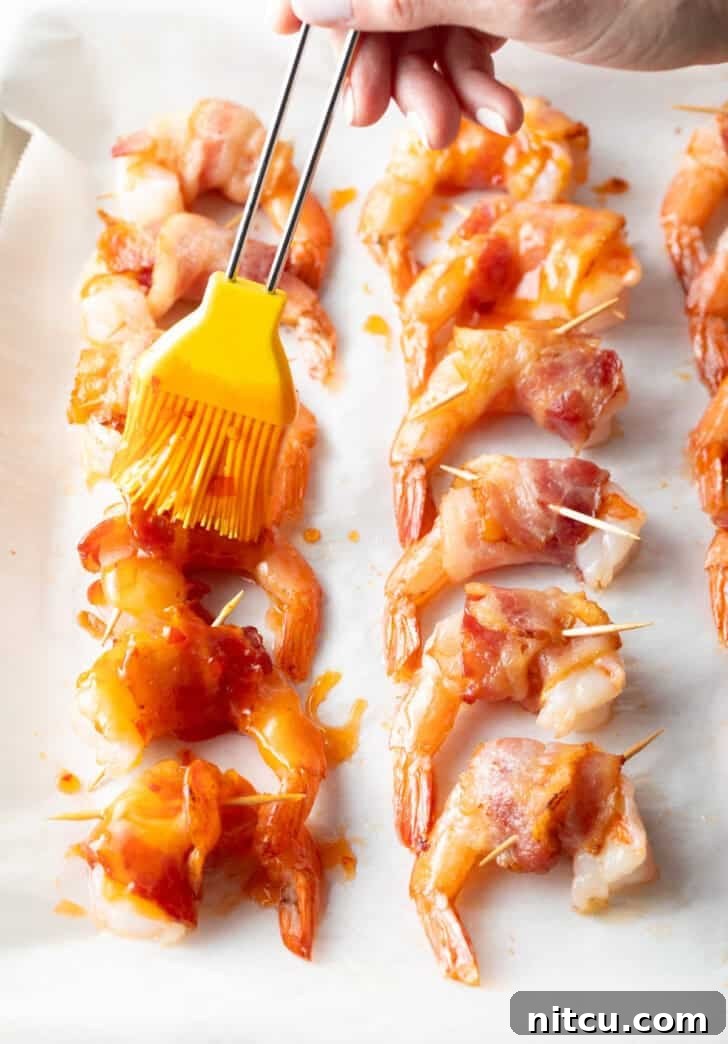 A vibrant yellow silicone brush expertly applies a glistening layer of Thai sweet chili sauce to a dozen bacon-wrapped shrimp, neatly arranged on a baking sheet, enhancing their flavor and adding a beautiful sheen.