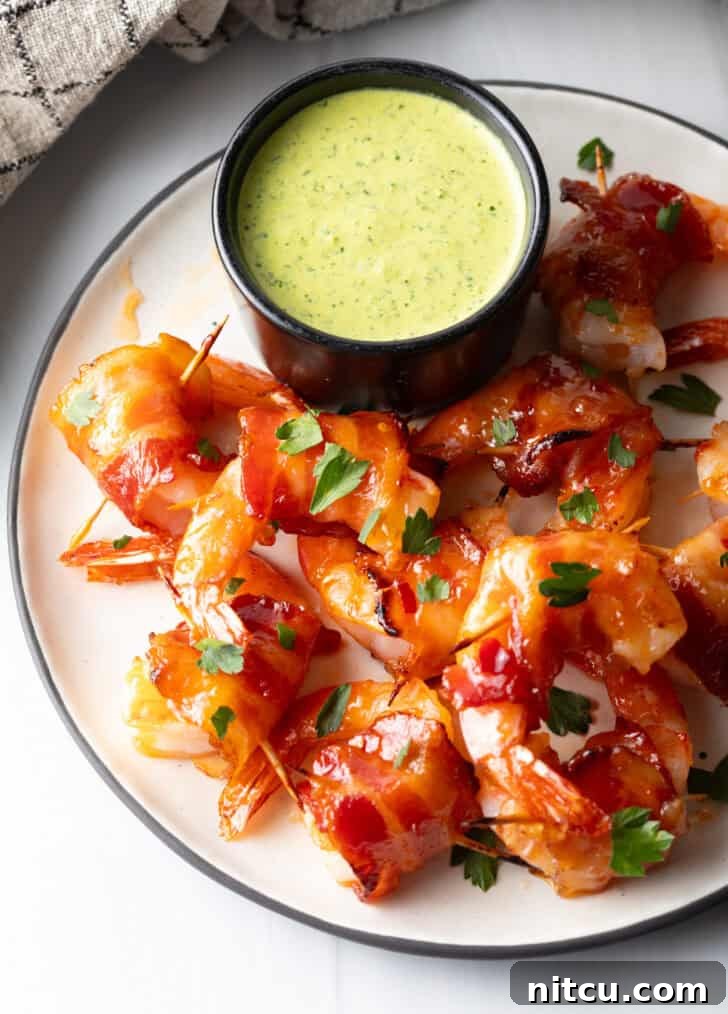 A white plate beautifully presents a black ramekin overflowing with vibrant green sauce, encircled by a plentiful serving of golden-brown bacon-wrapped shrimp, creating an inviting and colorful appetizer display.