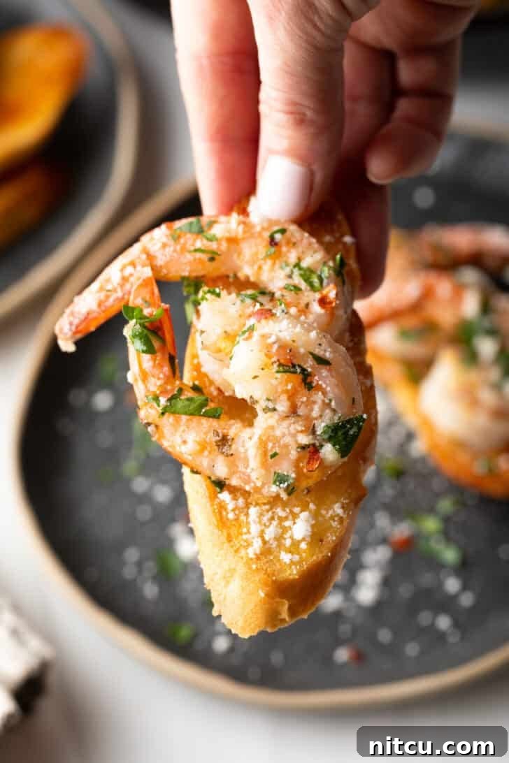 A hand holding a piece of toasted baguette generously topped with succulent garlic butter shrimp and fresh herbs, ready to be enjoyed.