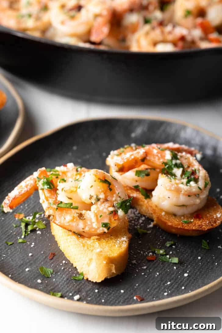 Two pieces of toasted baguette topped with generous portions of garlic butter shrimp and fresh herbs, presented on an elegant black oval plate.