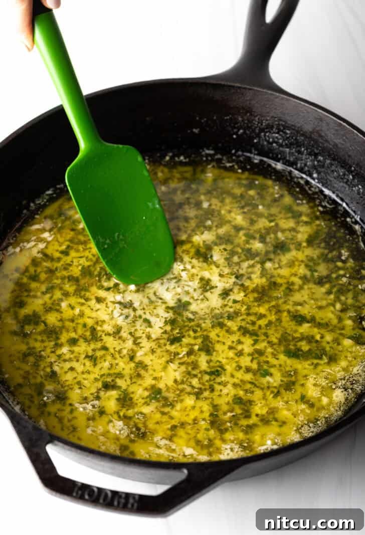 A green spatula stirring minced garlic, melted butter, and fresh herbs in a large skillet, creating an aromatic base for the shrimp scampi.