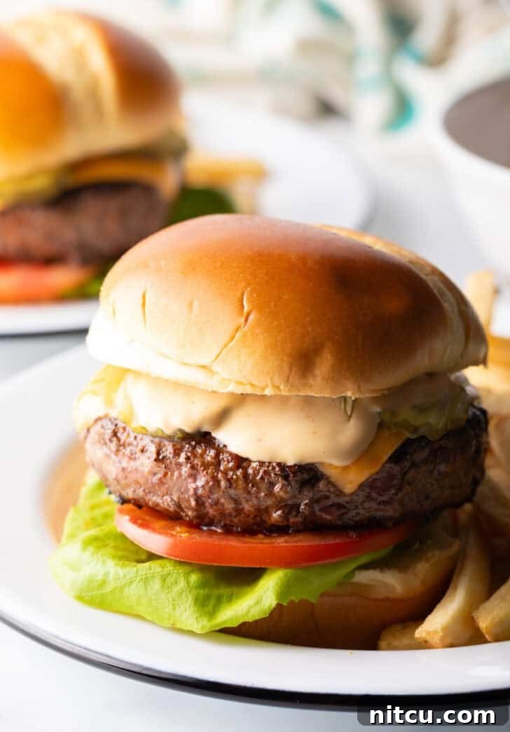 A delectable cheeseburger featuring lettuce, tomato, melted cheese, and a generous layer of homemade burger sauce, served alongside crispy french fries on a white plate.