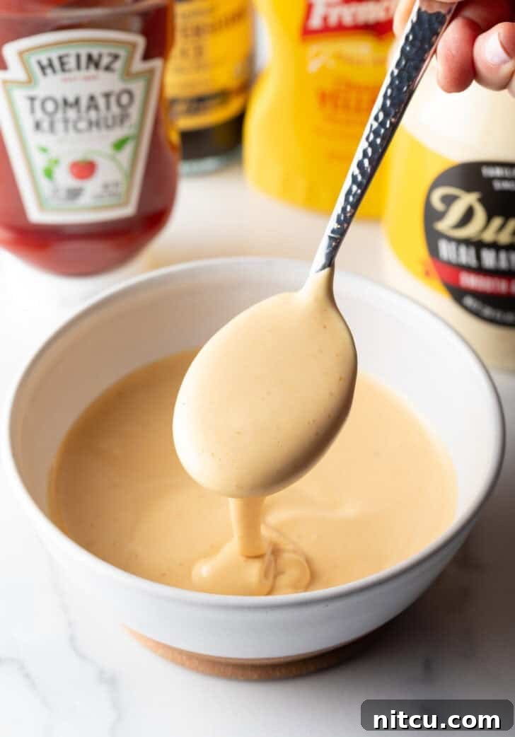 A spoonful of burger sauce is gracefully lifted over a bowl of the freshly made condiment, showcasing its creamy texture.