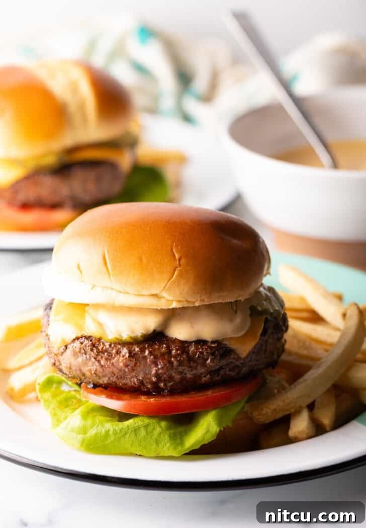 A mouthwatering cheeseburger, complete with fresh lettuce, ripe tomato slices, melted cheese, and a generous layer of homemade Burger King-style sauce. It's elegantly presented on a white plate with a side of golden french fries.