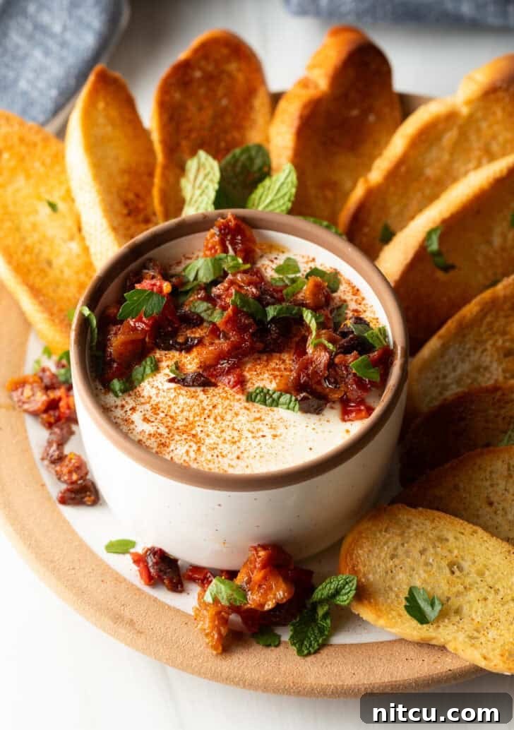 White cup with whipped feta with chopped sun dried tomato, on a plate surrounded by slices of toasted baguette.