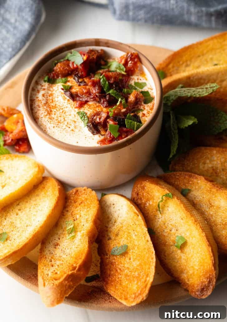 White cup with whipped feta with chopped sun dried tomato, on a plate surrounded by slices of toasted baguette.