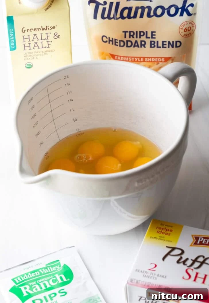 Recipe ingredients neatly arranged: a large white mixing pitcher containing several eggs, a box of puff pastry, a packet of ranch dressing mix, a bag of shredded cheddar cheese, and a carton of half and half.