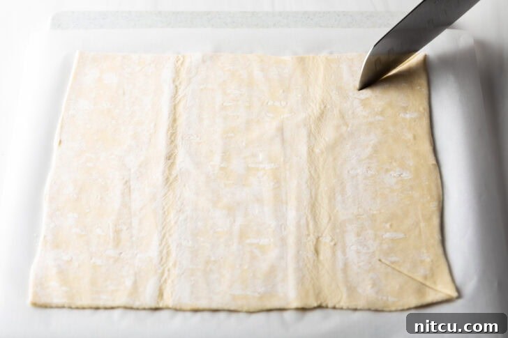 Using a sharp knife to carefully slice a large rectangle of puff pastry to the correct dimensions for the baking dish.