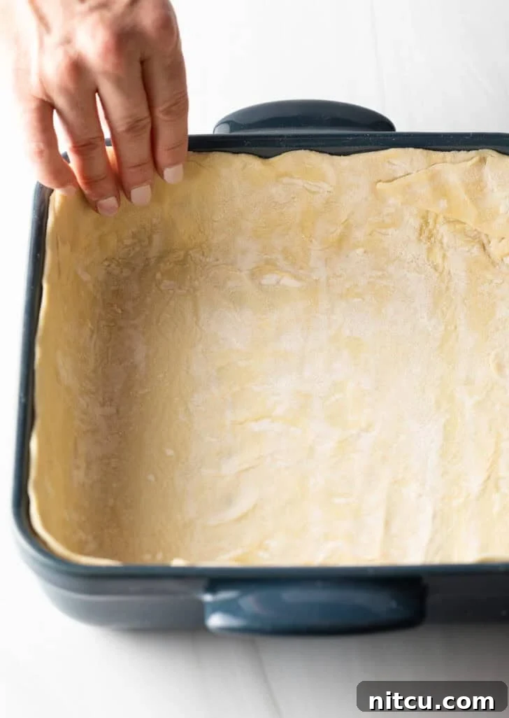 A skilled hand gently placing the prepared puff pastry dough into the bottom and up the sides of a square baking dish, creating a perfect crust.