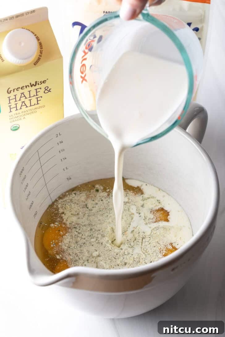 Pouring creamy half and half from a clear glass measuring cup into a large white mixing pitcher already filled with cracked eggs, ready for whisking.