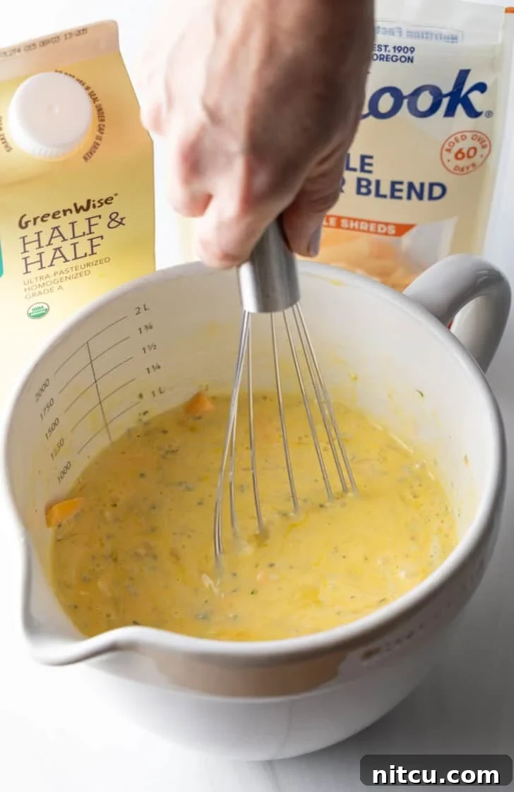 A hand vigorously whisking eggs, half and half, and shredded cheese together in a large white mixing pitcher to create a smooth, well-combined egg mixture.