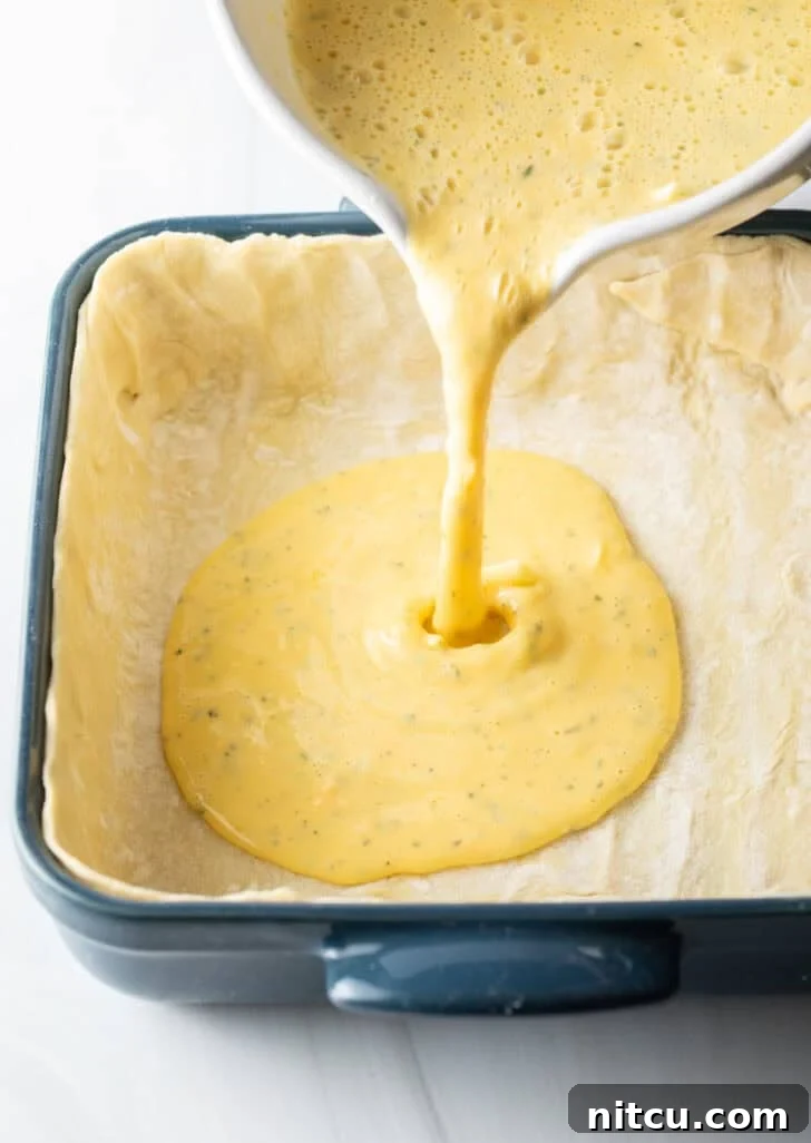 Carefully pouring the perfectly blended egg mixture from a measuring cup into a prepared square baking dish, already lined with the delicate puff pastry crust.