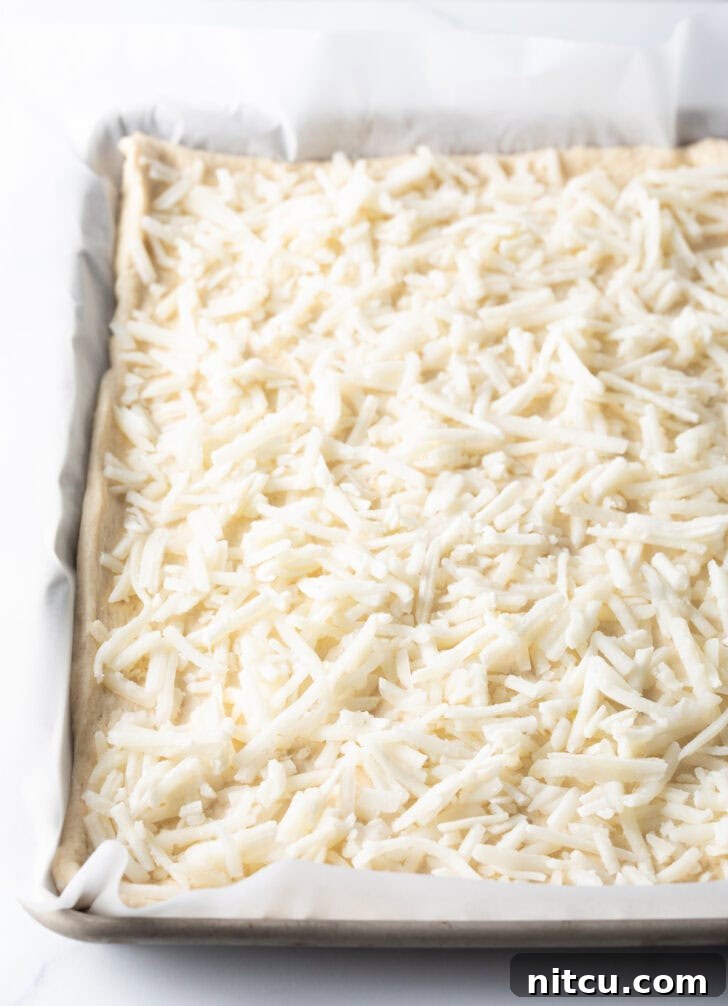 A close-up shot of an unbaked rectangular pizza crust, generously topped with a layer of shredded mozzarella cheese, ready for the oven.