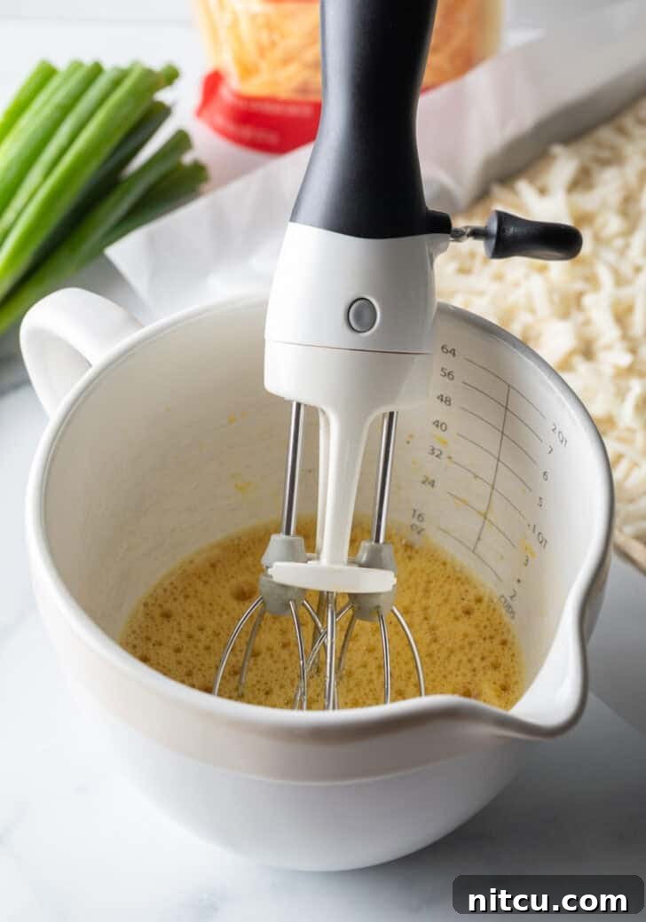 A white mixing pitcher containing a whisked egg mixture, with a hand holding a hand mixer, illustrating the preparation process for the fluffy eggs.