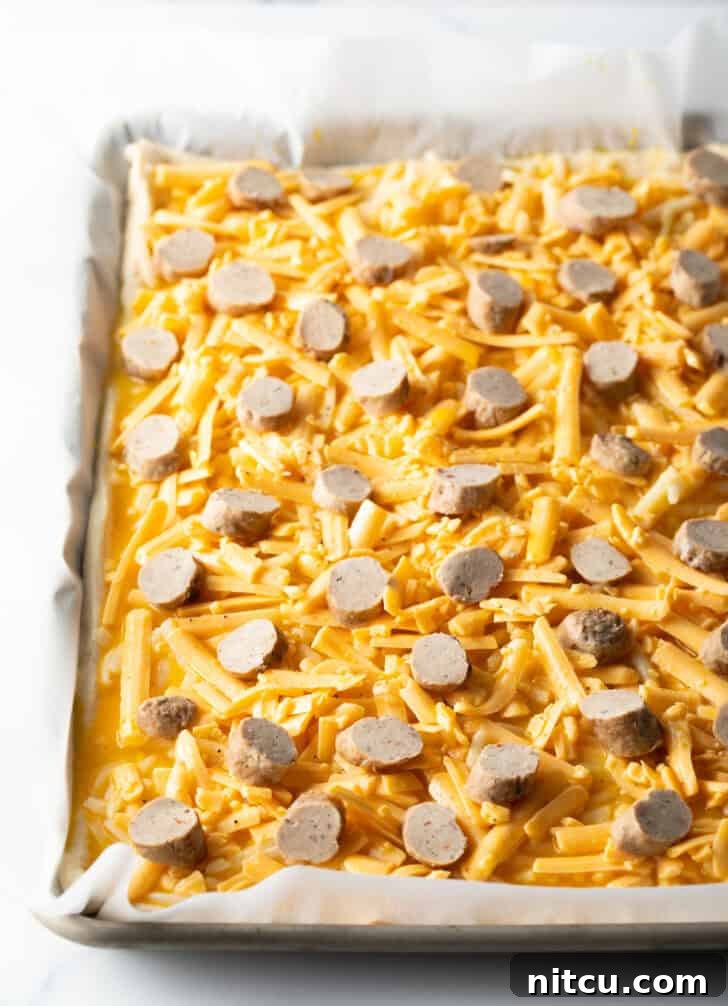 An unbaked rectangular pizza base layered with shredded mozzarella cheese and evenly distributed slices of breakfast sausage, poised for baking.