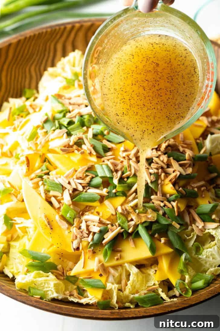 Pouring dressing from a glass measuring cup into a bowl with shredded cabbage, toasted almonds, green onion slices, and slices of mango.