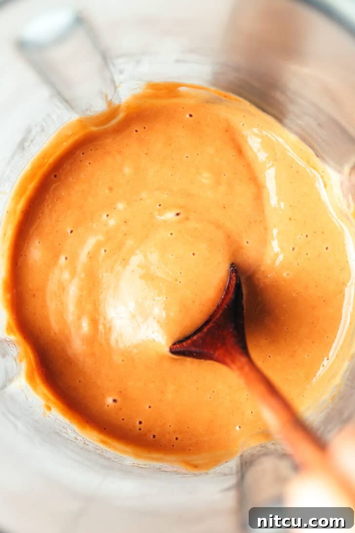 A wooden spoon actively stirring the creamy peanut sauce inside a blender jar, ensuring a smooth and perfectly combined consistency.