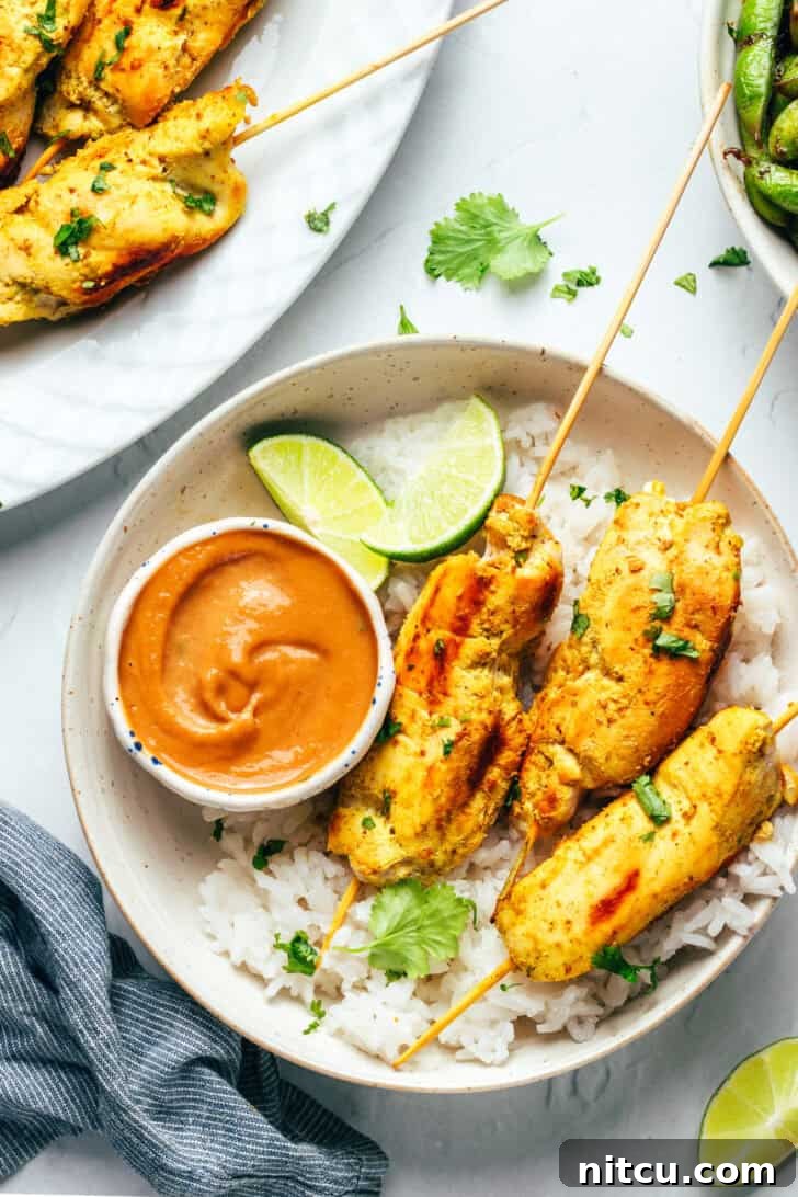 Six pieces of chicken satay arranged on a white round plate with a side of fluffy white rice, vibrant lime slices, and a small white ramekin of rich peanut sauce.